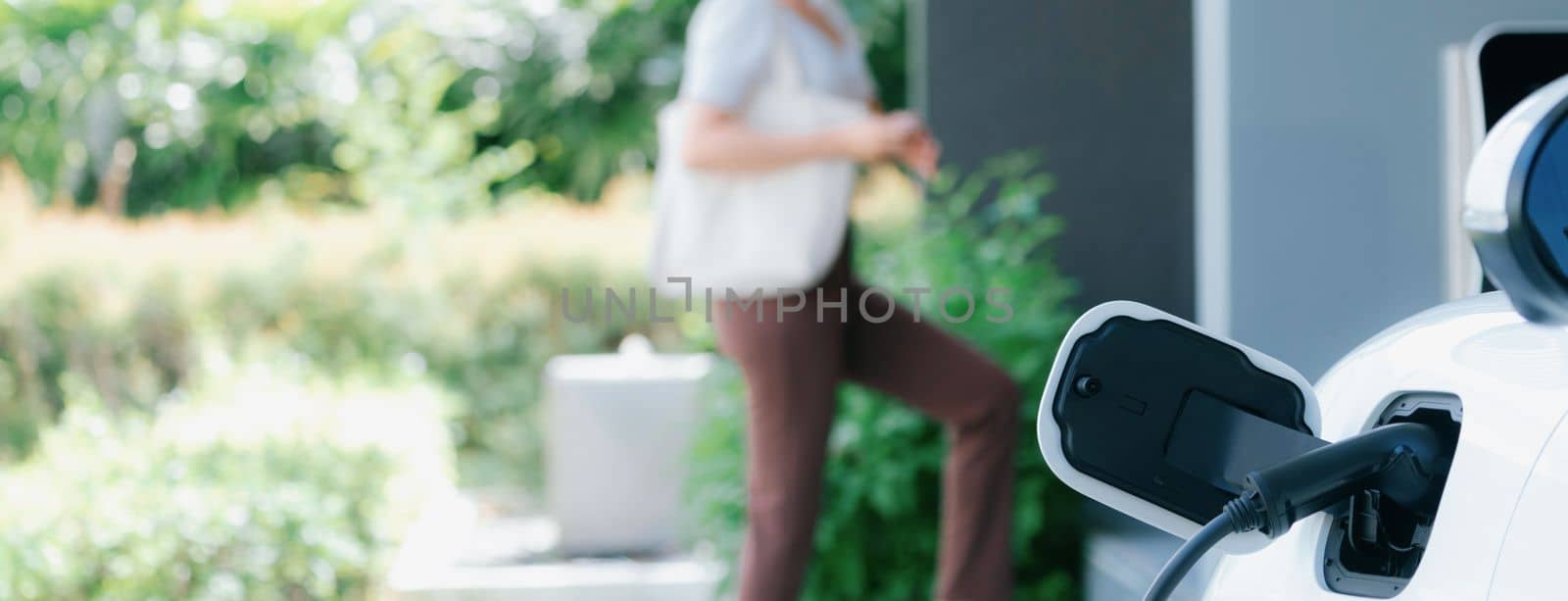 Focus charger plug into EV car with blur backdrop of progressive woman walking. by biancoblue