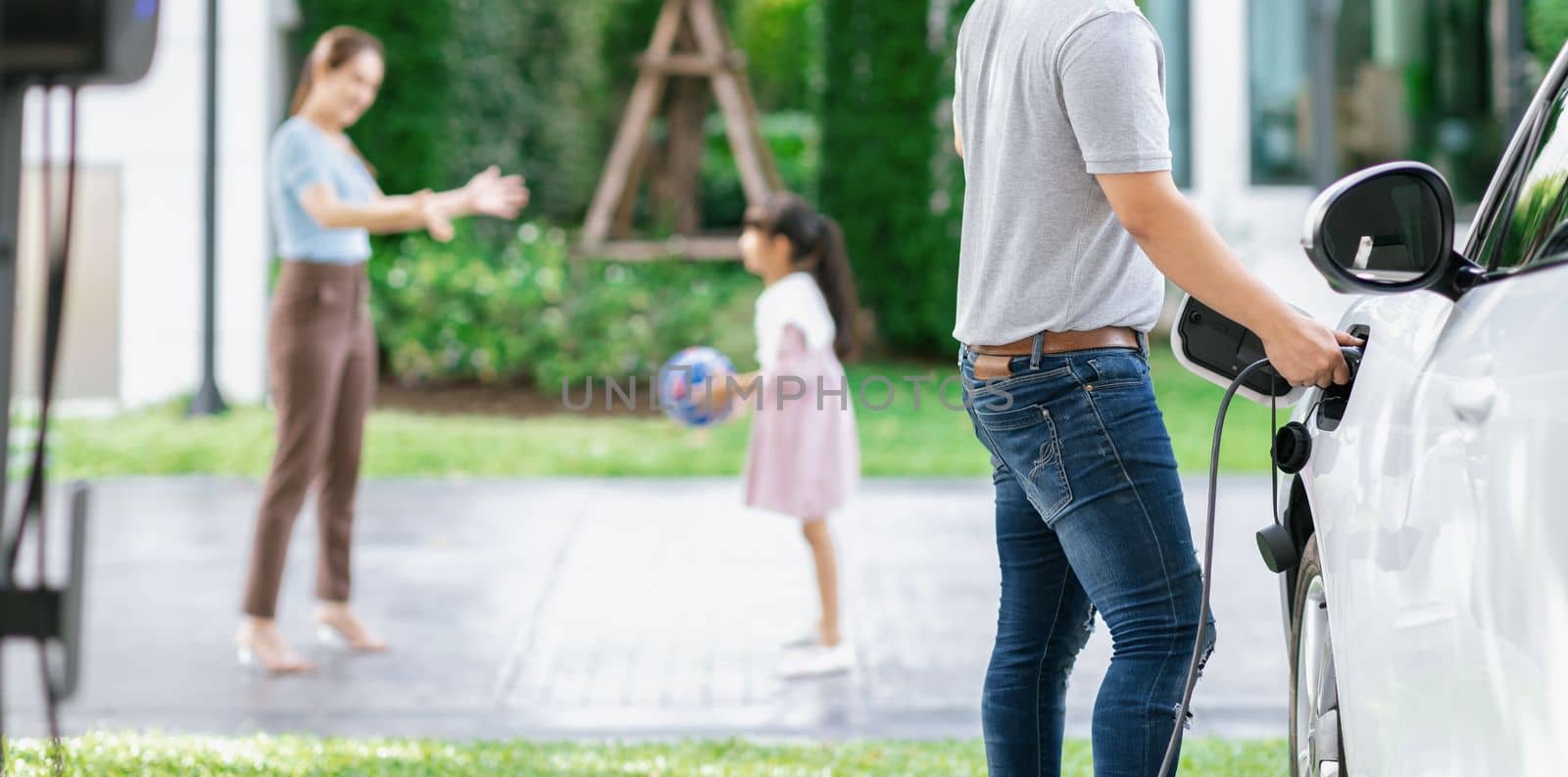 Focus image of progressive man charging electric car from home charging station with blur mother and daughter playing together in the background.