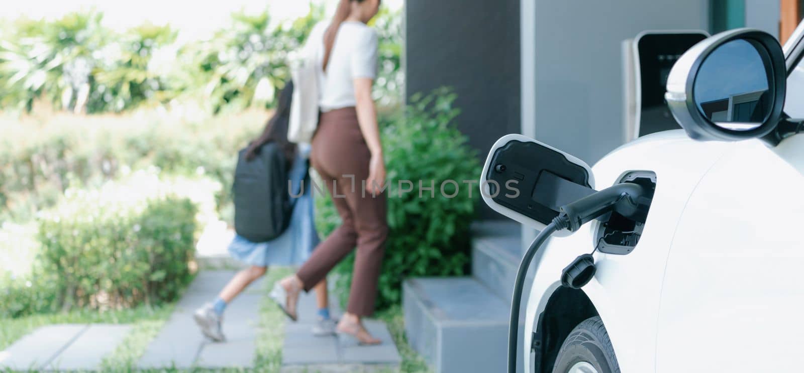 Focus electric vehicle recharging at home charging station plugged in with EV charger device with blurred background of progressive mother and daughter walking as concept for sustainability of energy.