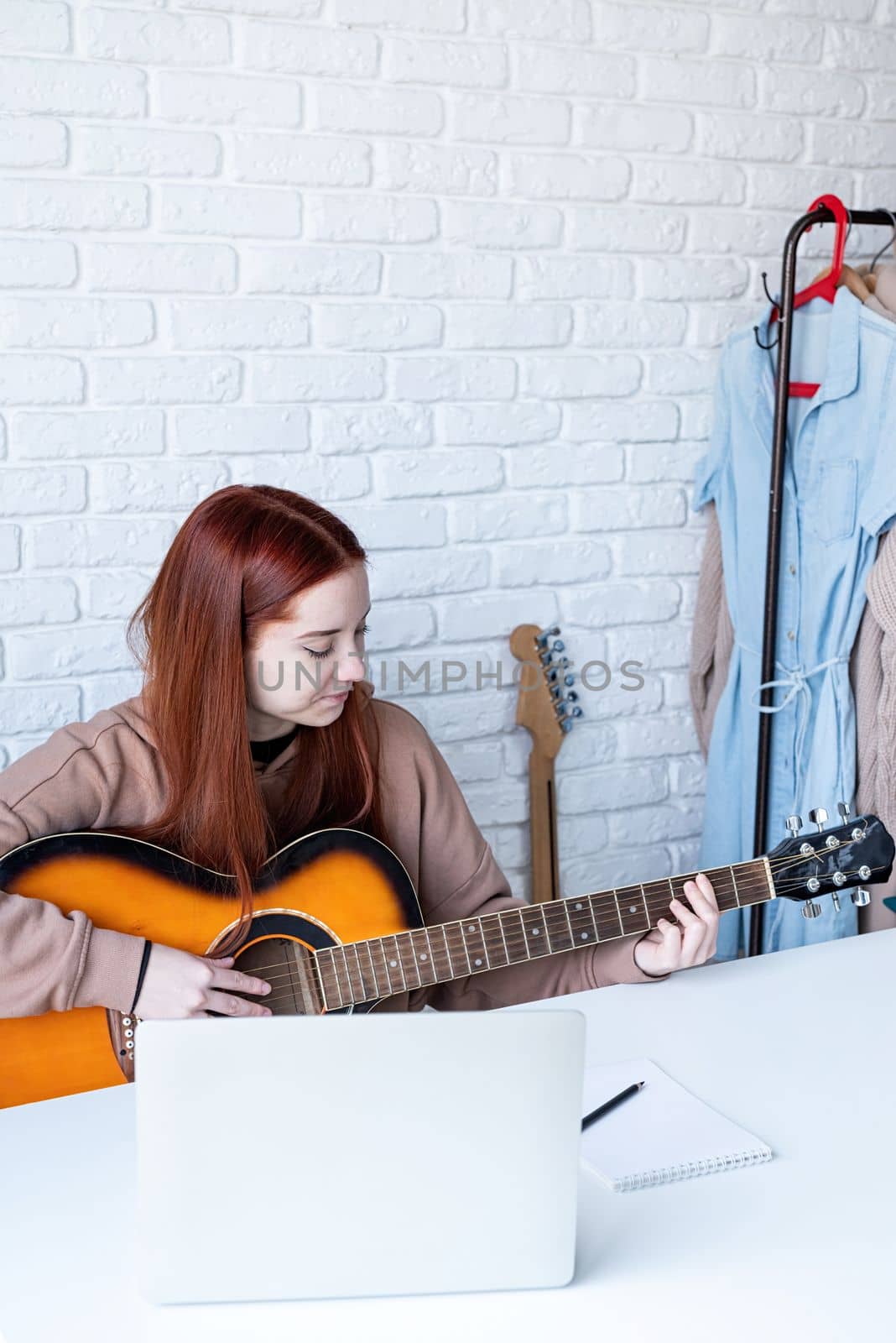 young caucasian red-haired woman learning to play acoustic guitar at home, making notes and using laptop
