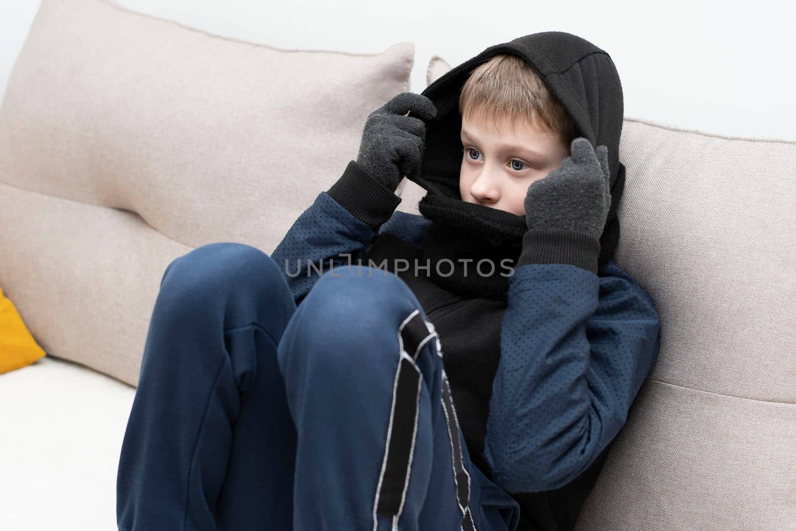 People. Energy crisis concept. A teenage boy sits at home on the sofa in gloves, a warm scarf and a hood on his head, freezes due to the heating being turned off. War in Europe. Cold in apartments. Energy crisis Ukraine. Close-up.
