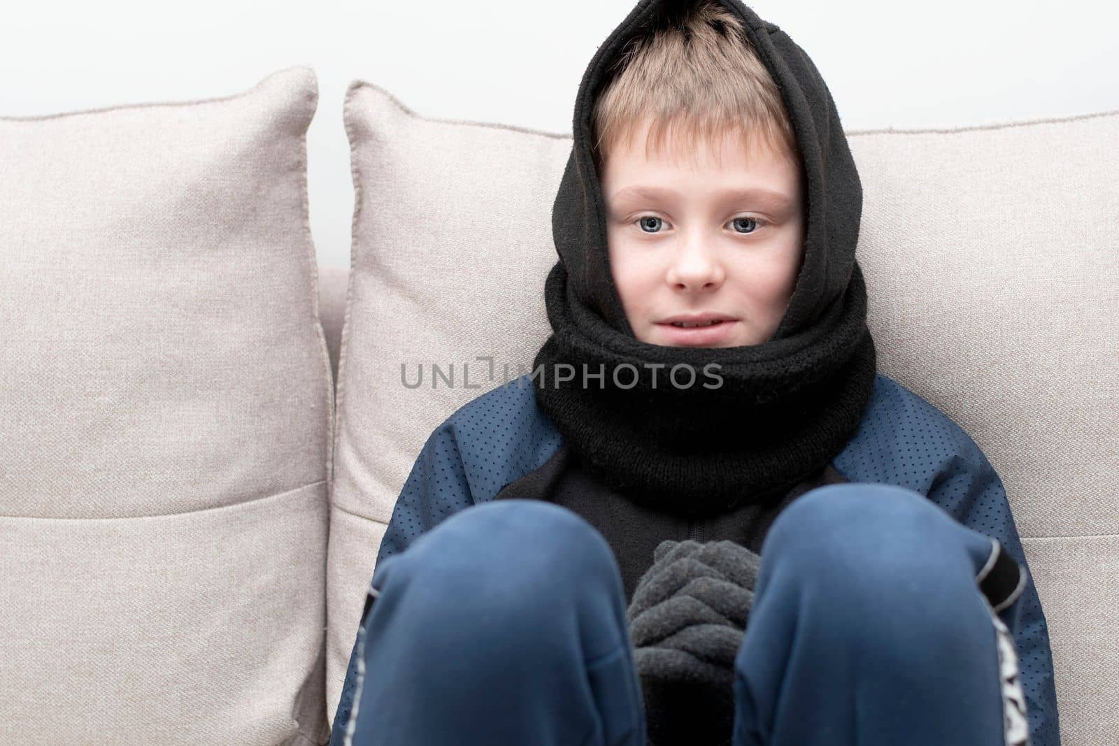 People. Energy crisis concept. A teenage boy sits at home on the sofa in gloves, a warm scarf and a hood on his head, freezes due to the heating being turned off. War in Europe. Cold in apartments. Energy crisis Ukraine. Close-up.