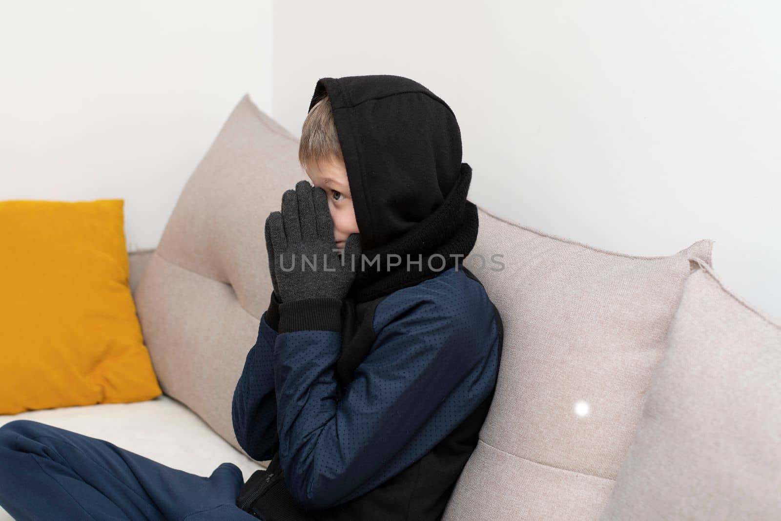 People. Energy crisis concept. A teenage boy sits at home on the sofa in gloves, a warm scarf and a hood on his head, freezes due to the heating being turned off. War in Europe. Cold in apartments. Energy crisis Ukraine. Close-up.