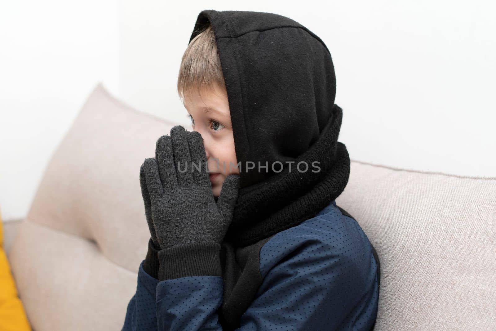 People. Energy crisis concept. A teenage boy sits at home on the sofa in gloves, a warm scarf and a hood on his head, freezes due to the heating being turned off. War in Europe. Cold in apartments. Energy crisis Ukraine. Close-up.