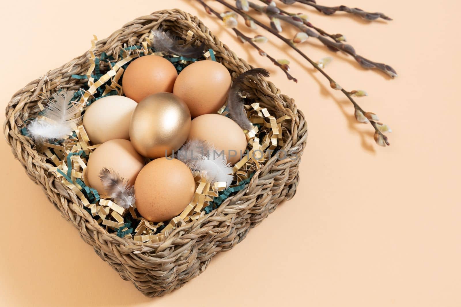 Easter eggs natural and gold in basket with feather. Branch of willow catkins. Light pastel orange background with copy space.