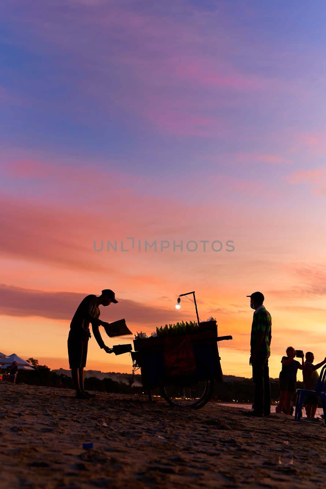 An evening silhouette of a vendor and his grill cart on the beach. Evening market on the beach. Bali, Indonesia - 11.29.2022