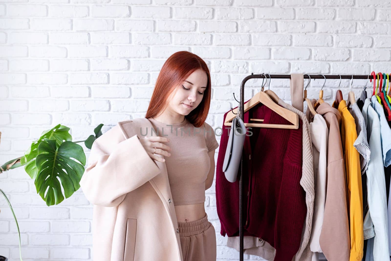 young beautiful woman wearing beige pastel coat, standing near clothes rack. Wardrobe change by Desperada