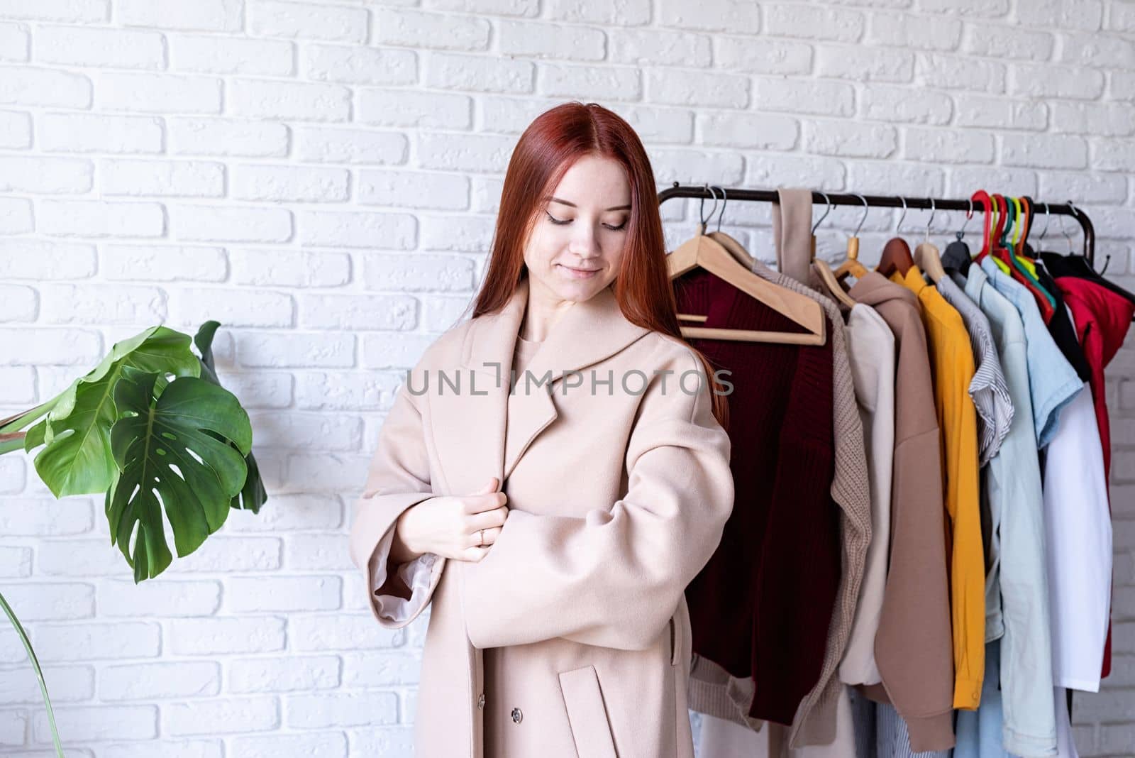 young beautiful woman wearing beige pastel coat, standing near clothes rack. Wardrobe change by Desperada
