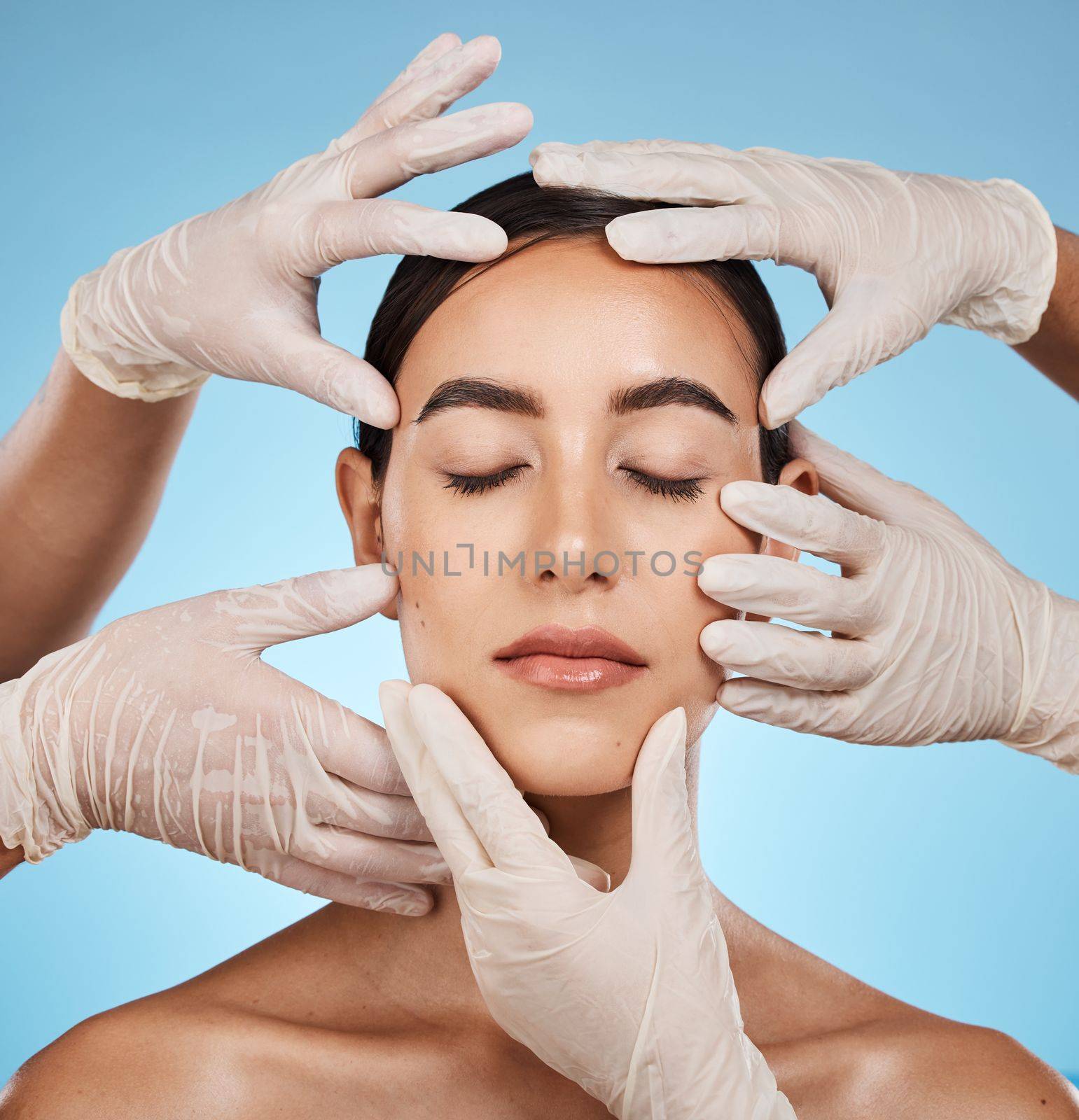 Plastic surgery, beauty and hands on the face of a woman isolated on a blue background in a studio. Feeling, skincare and doctors touching a model for a botox, cosmetics or dermatology consultation by YuriArcurs