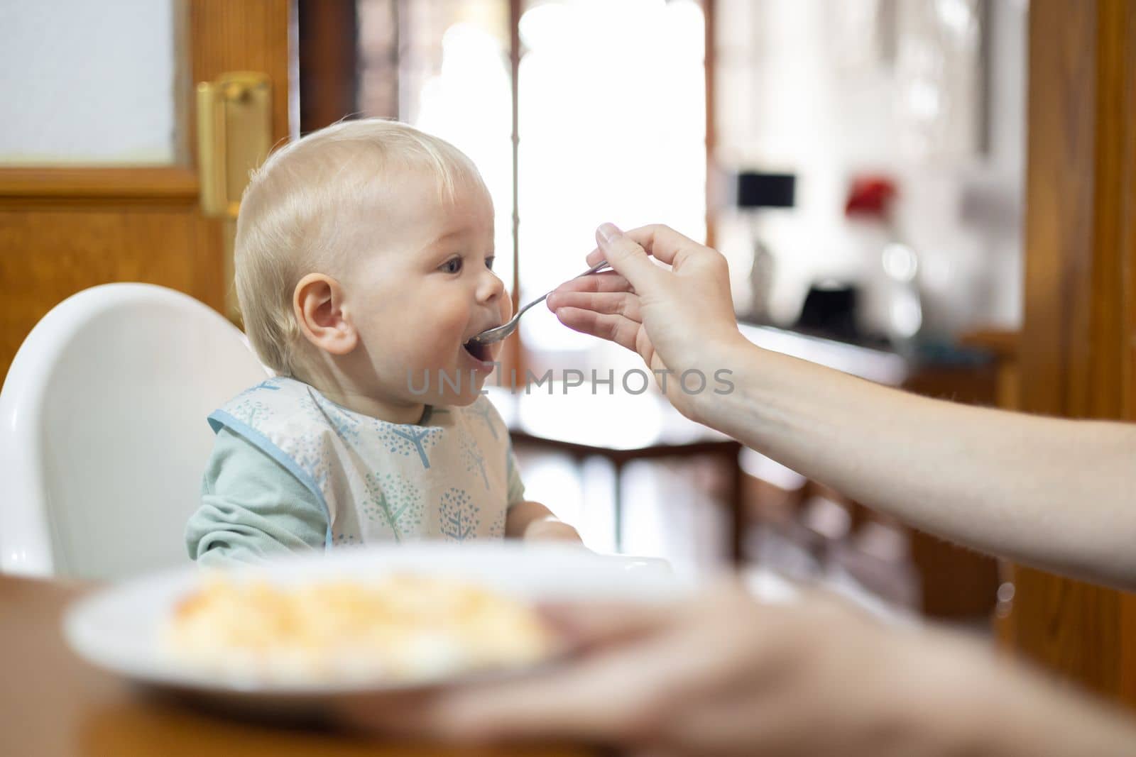 Mother spoon feeding her infant baby boy child sitting in high chair at the dining table in kitchen at home.