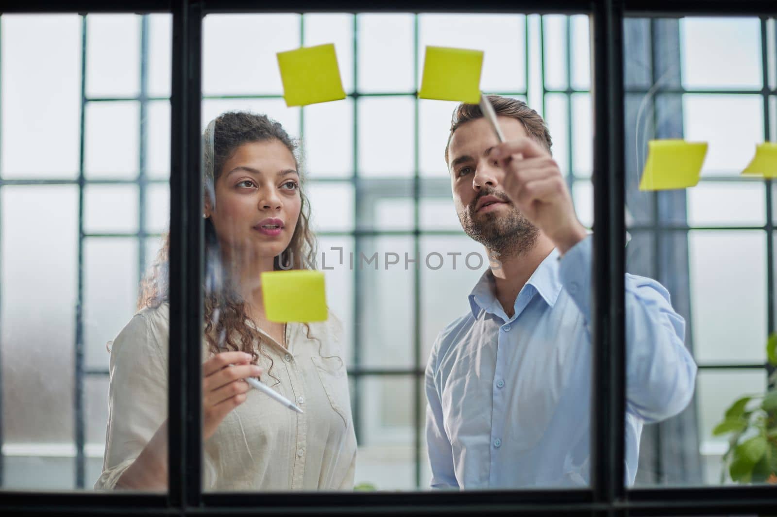 Creative professionals standing and discussing in office behind glass wall with sticky notes