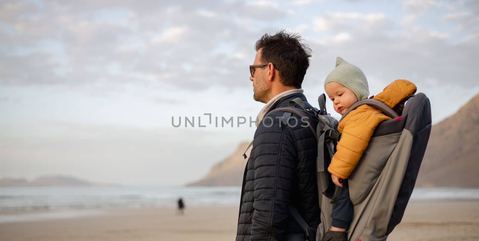 Father enjoying pure nature carrying his infant baby boy son in backpack on windy sandy beach of Famara, Lanzarote island, Spain at sunset. Family travel concept. by kasto