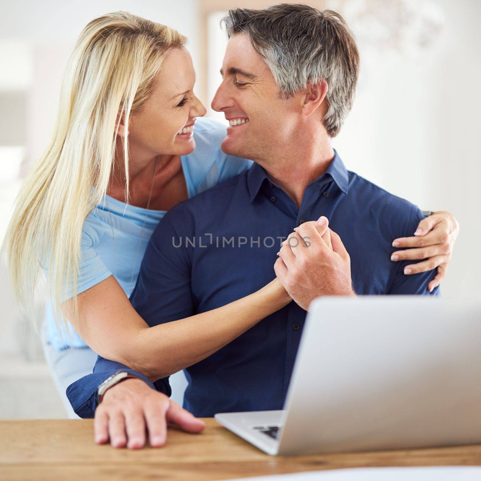 Together forever. an attractive mature woman hugging her handsome mature husband while he uses a laptop in the house. by YuriArcurs