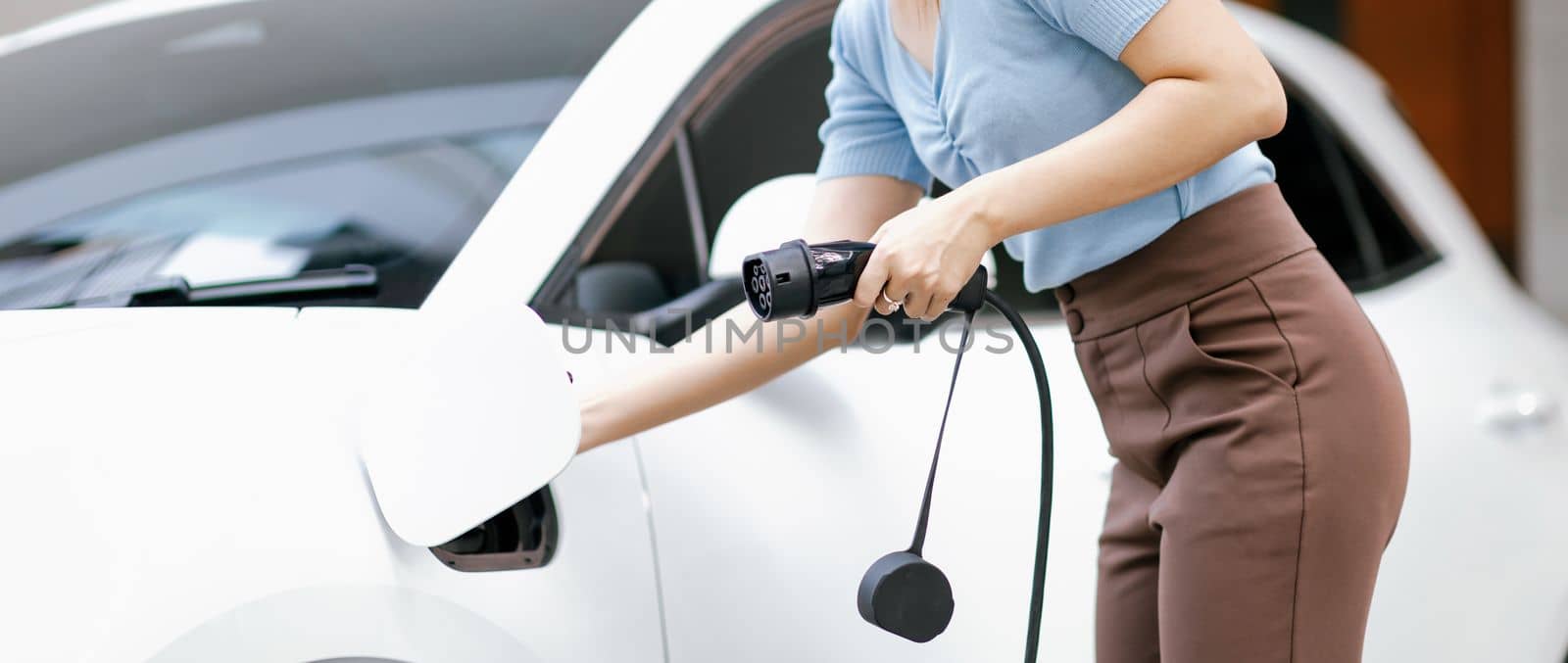 Closeup progressive woman install cable plug to her electric car with home charging station. Concept of the use of electric vehicles in a progressive lifestyle contributes to clean environment.