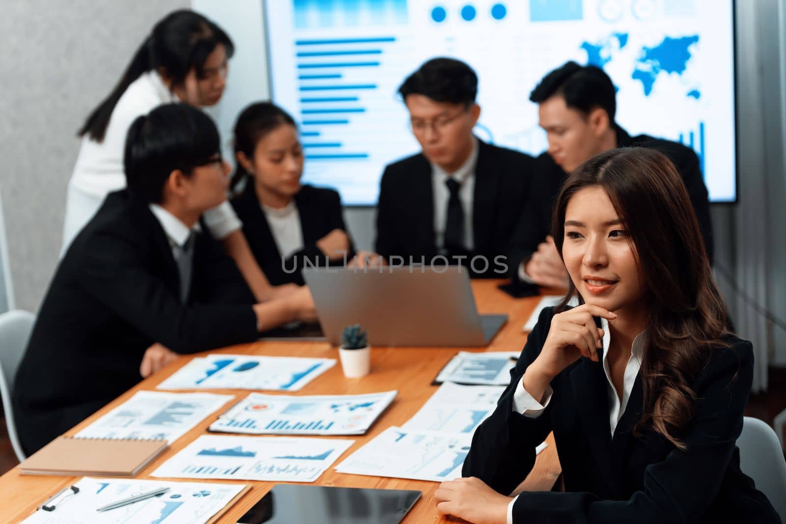 Focus portrait of female manger, businesswoman in the harmony meeting room with blurred of colleagues working together, analyzing financial paper report and dashboard data on screen in background.