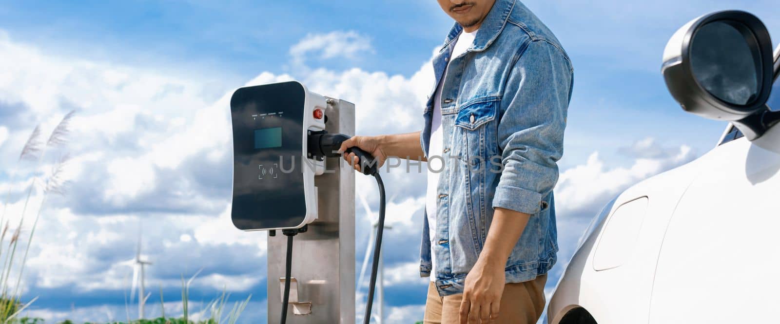 Progressive man with plugged-in EV car with charging point, cloudscape backdrop. by biancoblue