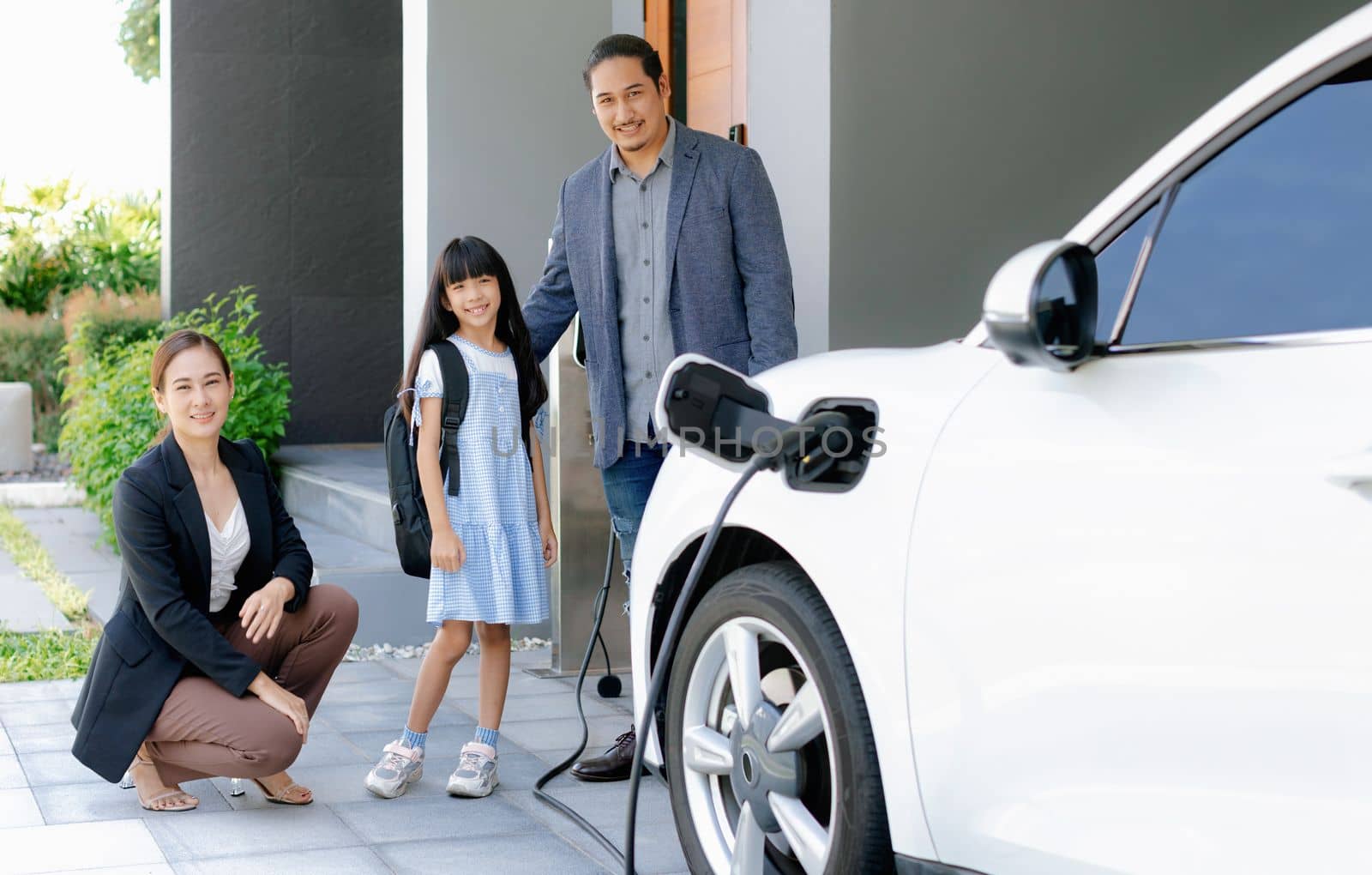 Progressive young parents and daughter with electric vehicle and home charging station. Green and clean energy from electric vehicles for healthy environment. Eco power from renewable source at home.