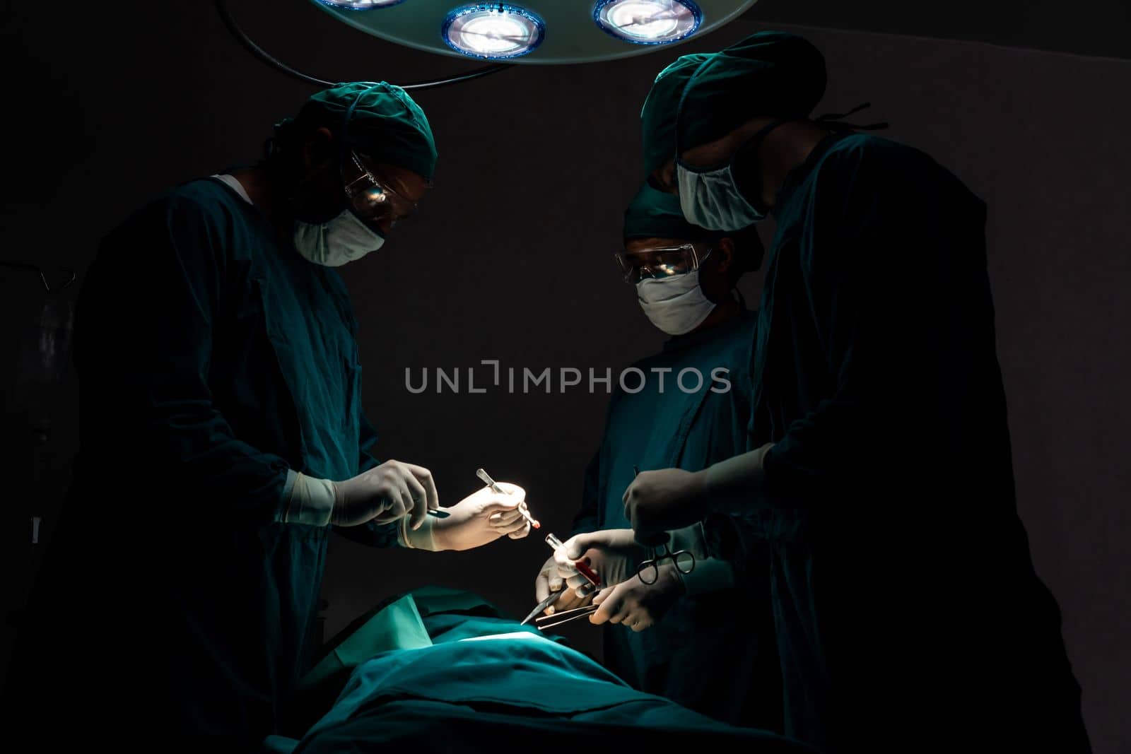 Surgical team performing surgery to patient in sterile operating room. In a surgery room lit by a lamp, a professional and confident surgical team provides medical care to an unconscious patient.