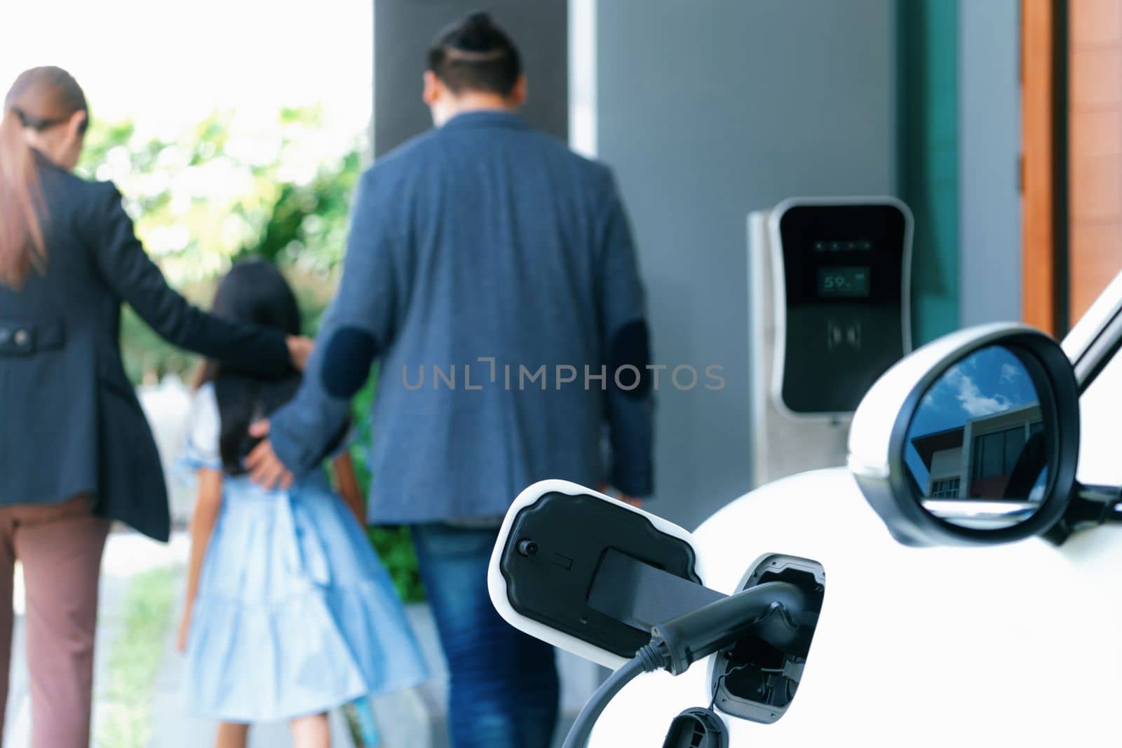 Focus closeup electric vehicle recharging battery from home electric charging station with blurred family in background. Renewable clean energy car for progressive eco awareness lifestyle concept.