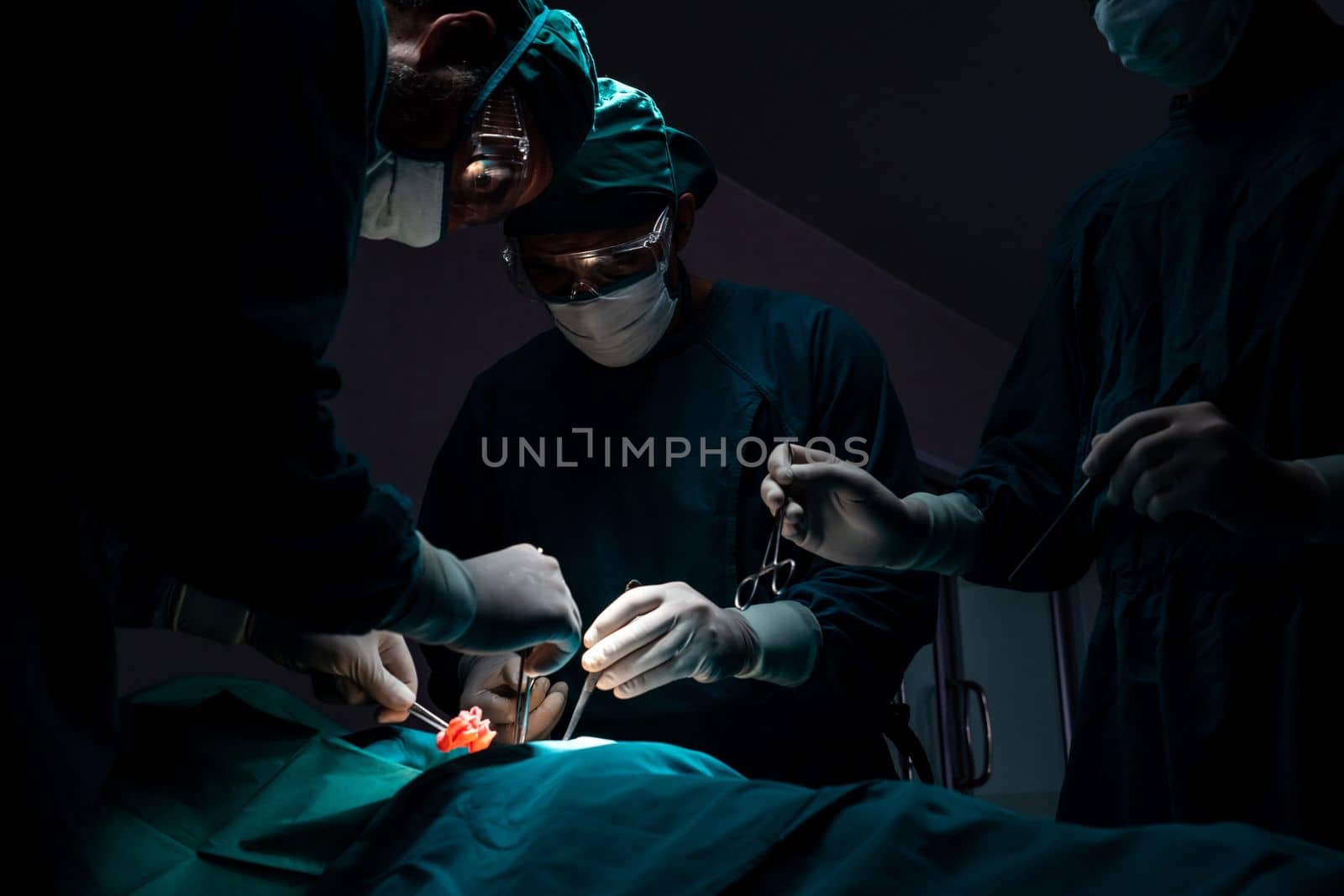 Surgical team performing surgery to patient in sterile operating room. In a surgery room lit by a lamp, a professional and confident surgical team provides medical care to an unconscious patient.