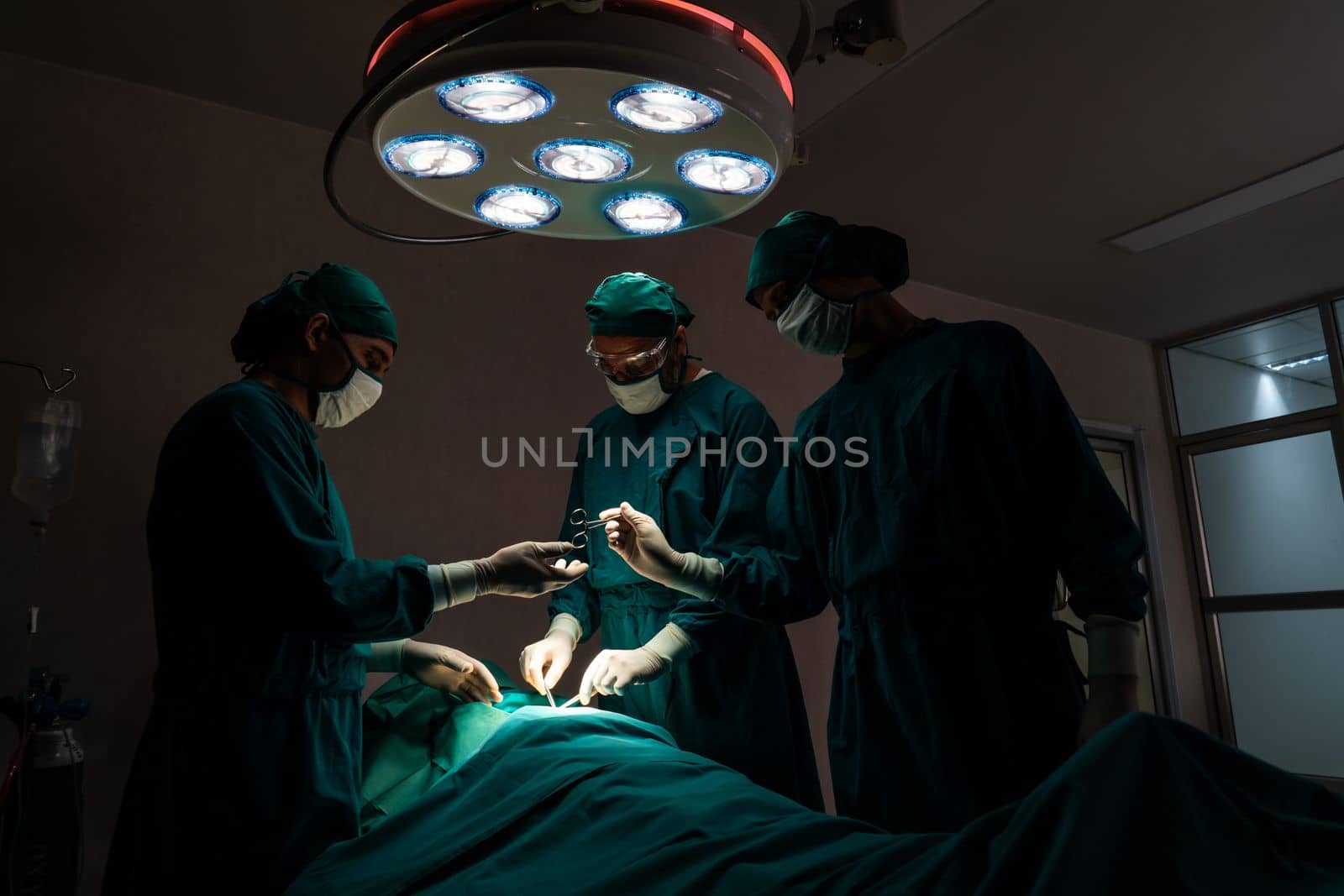 Surgical team performing surgery to patient in sterile operating room. In a surgery room lit by a lamp, a professional and confident surgical team provides medical care to an unconscious patient.