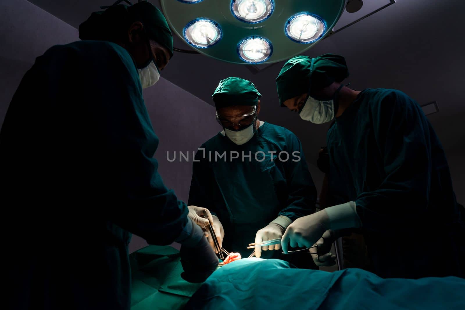 Surgical team performing surgery to patient in sterile operating room. In a surgery room lit by a lamp, a professional and confident surgical team provides medical care to an unconscious patient.