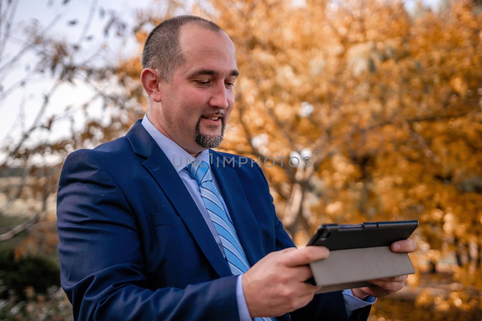 Mature business man in suit reading a news sitting in the park on the bench. Entrepreneur drink coffee in autumn park. Senior executive rest in city park. by panophotograph