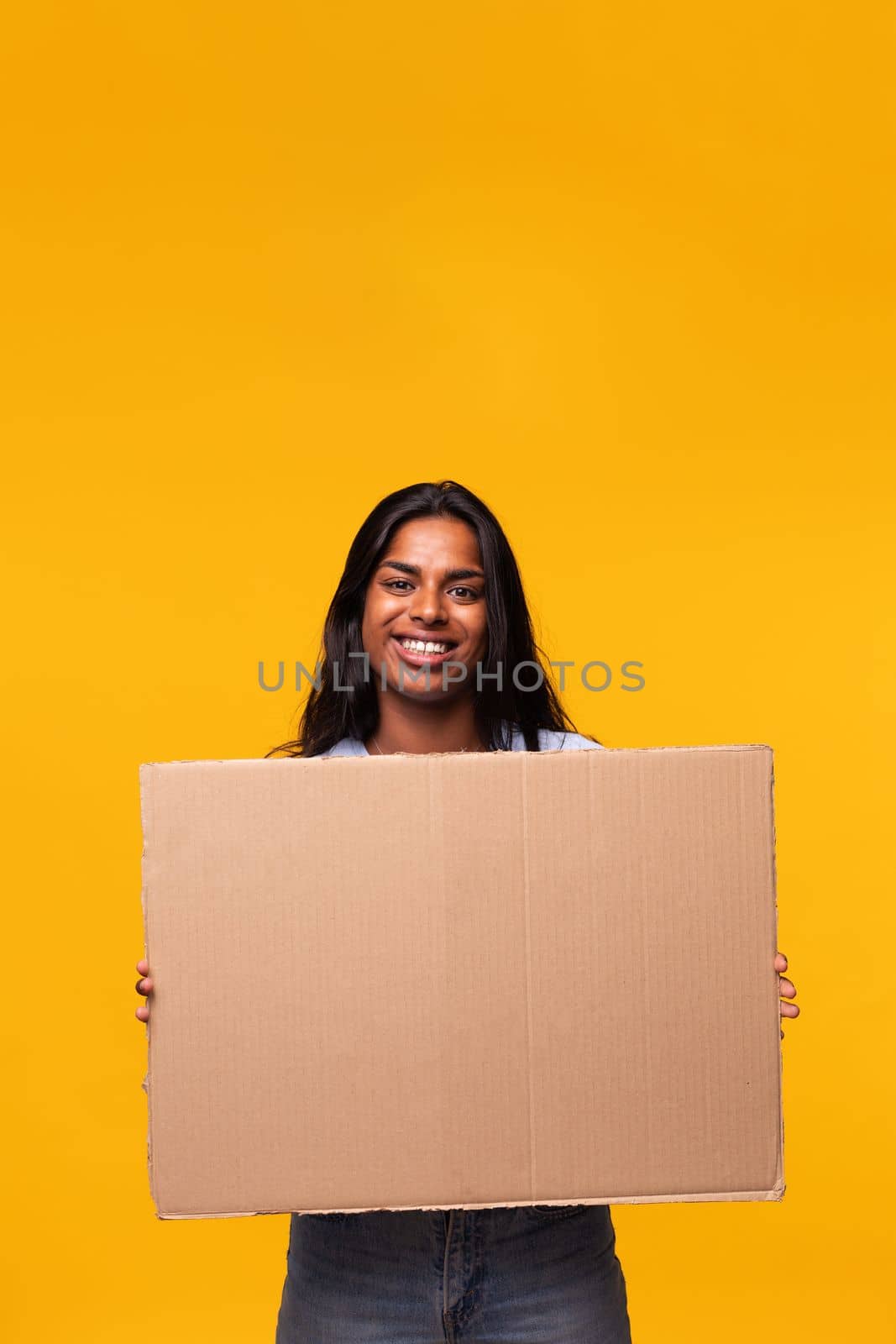 Young Indian asian woman looking at camera holding a cardboard banner isolated in yellow background. Studio shot. Vertical image.