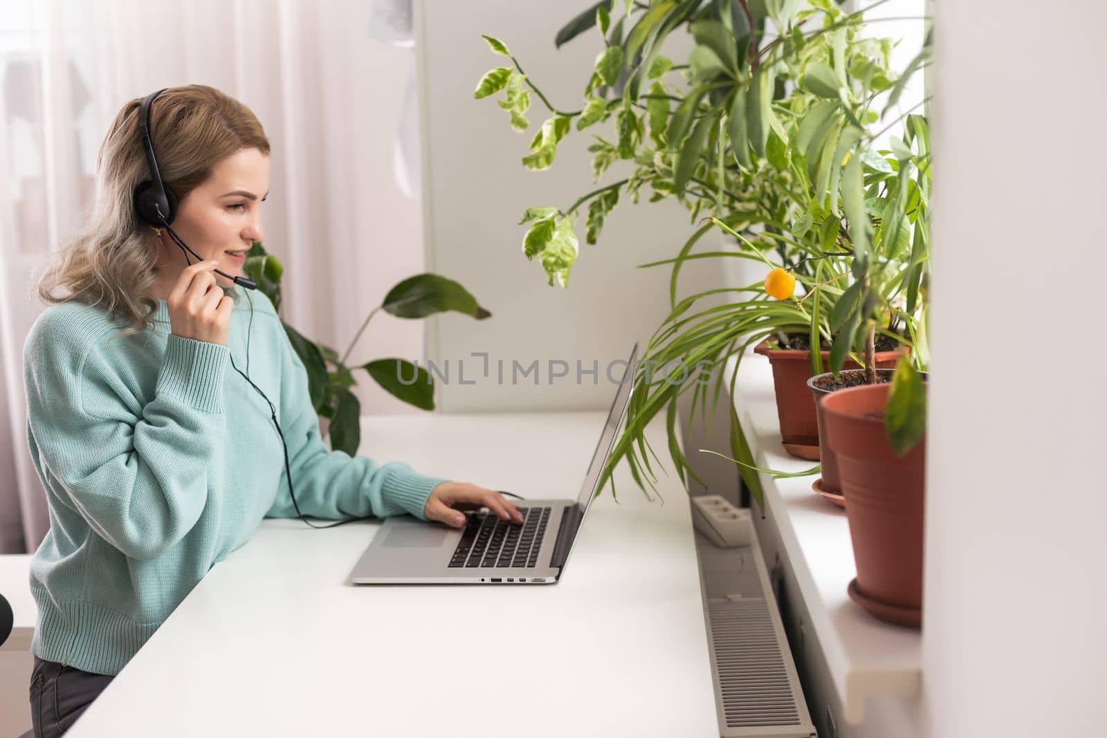 Positive young woman using a laptop at home, video conference. Cozy home interior with indoor plants. Remote work, business, freelance, online shopping, e-learning concept.