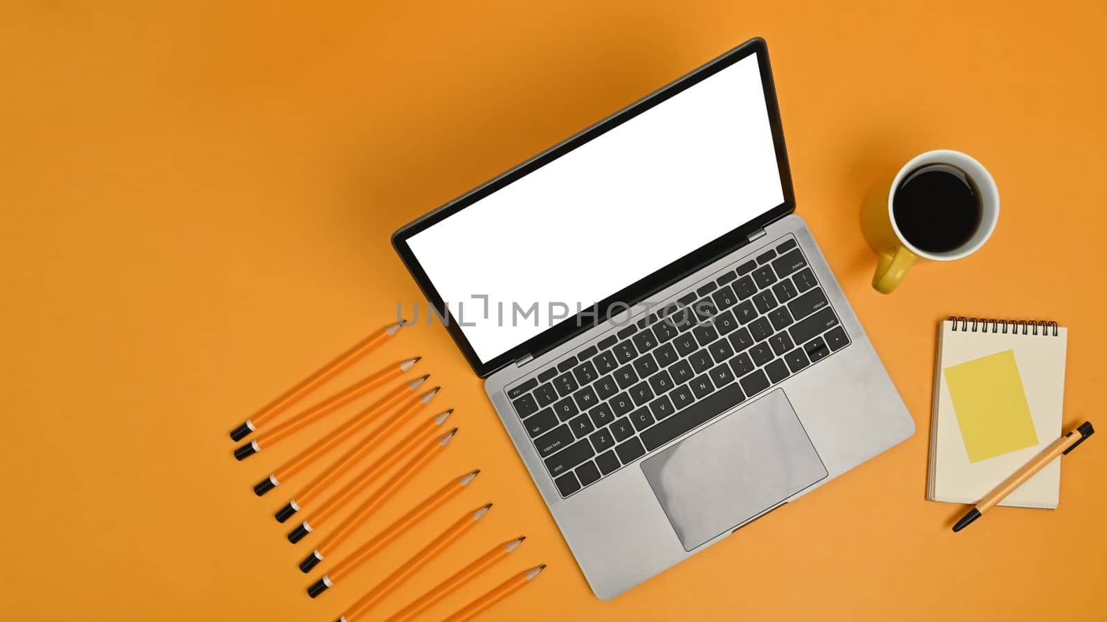 Mock up laptop computer with empire display, notepad, sticky note, pencils and coffee cup on yellow background.