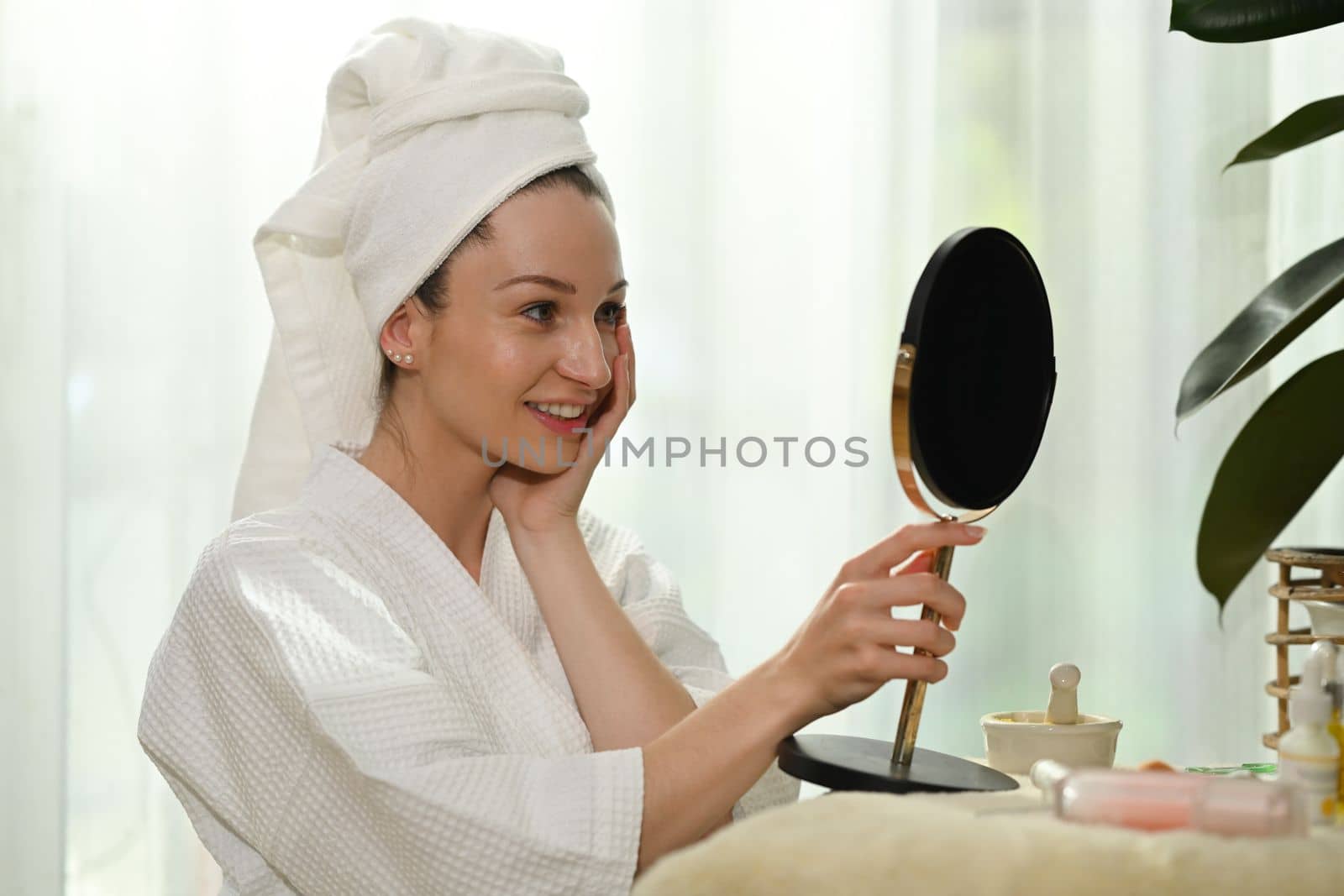 Smiling millennial woman in bathrobe standing front of mirror and making daily beauty routine at home.