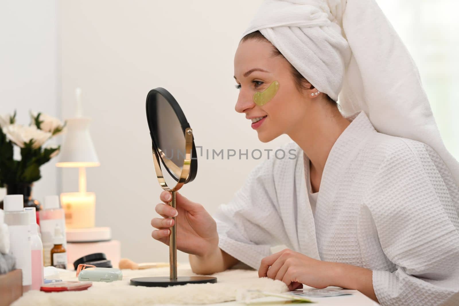 Beautiful caucasian woman applying anti fatigue under eyes and looking at mirror on dresser table. Beauty treatment concept.