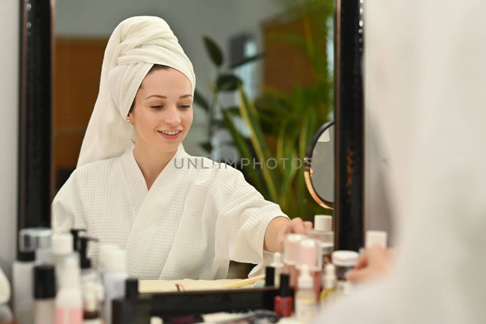 Millennial caucasian woman in bathrobe sitting front of mirror and applying cream, making daily beauty routine at home by prathanchorruangsak