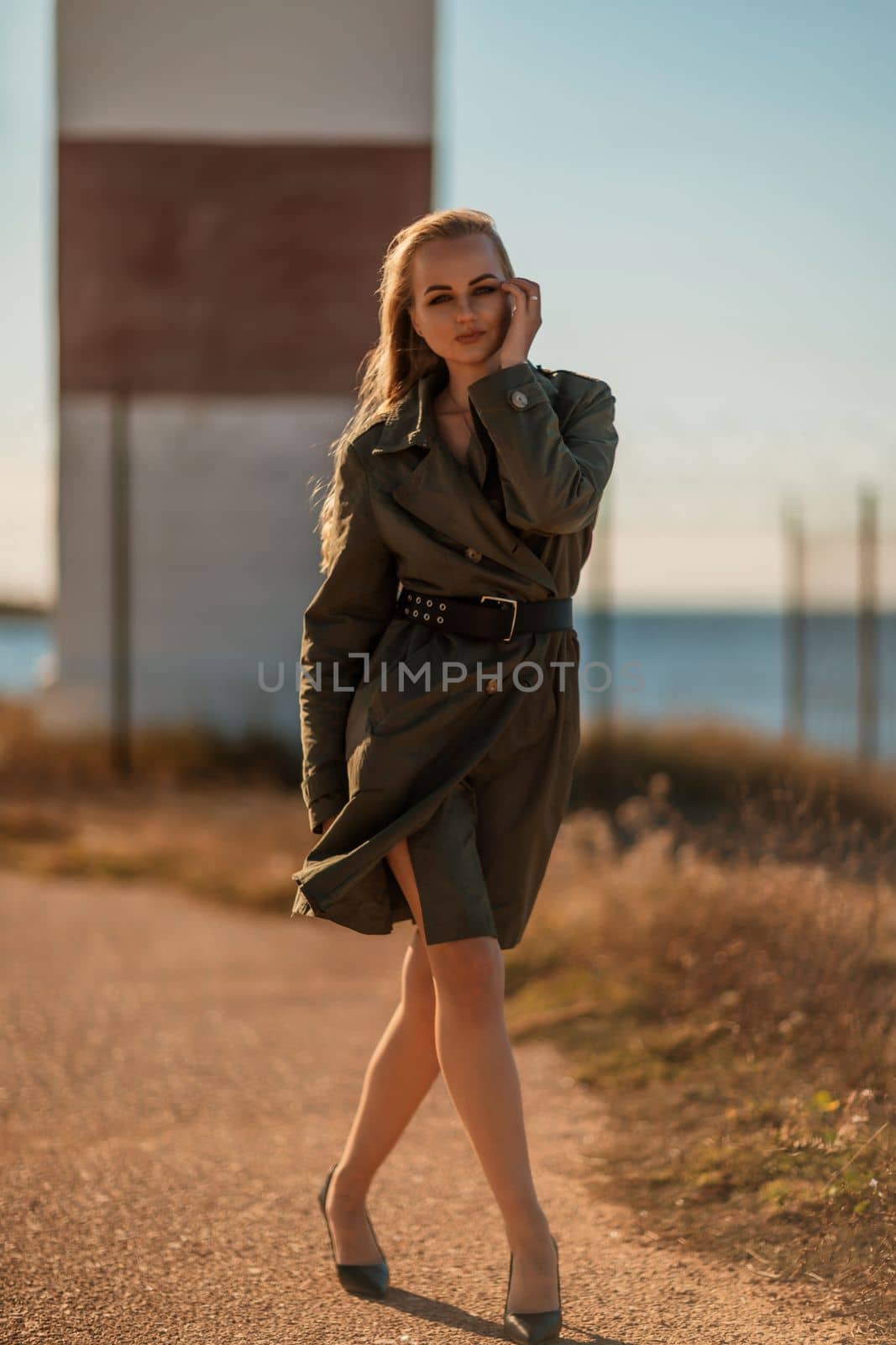 Portrait blonde sea cape. A calm young blonde in a khaki raincoat stands on the seashore against the backdrop of a lighthouse