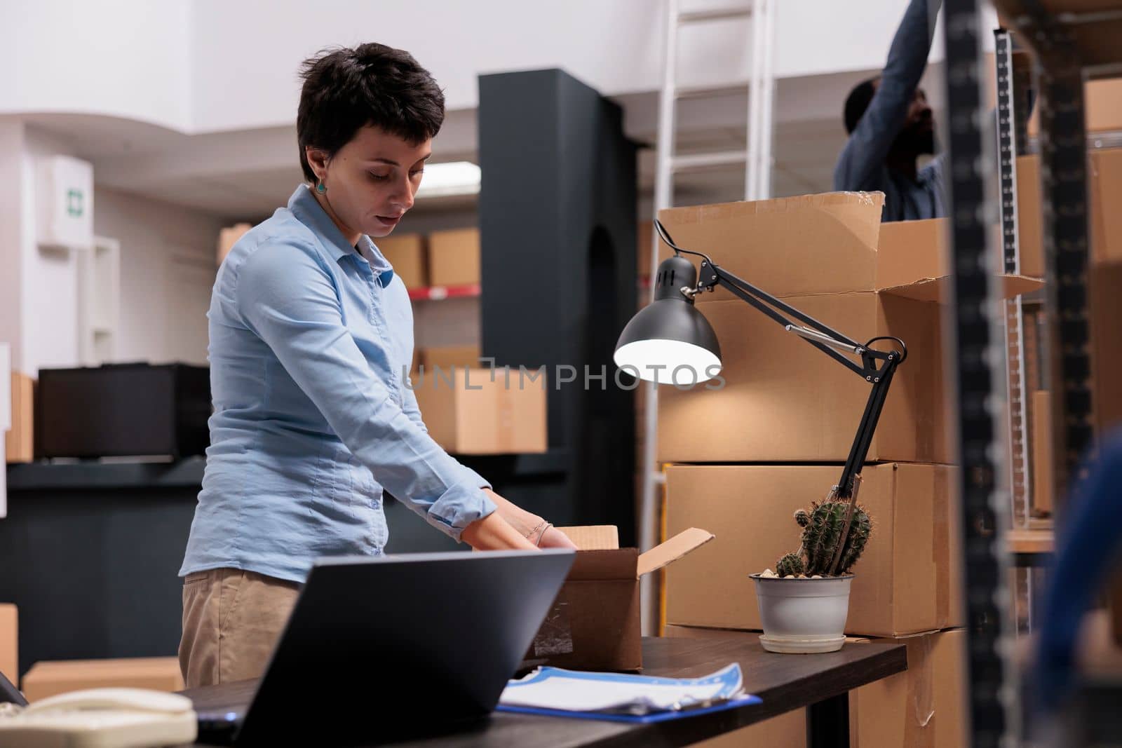 Caucasian manager preparing customers orders putting trendy clothes in carton boxes while looking at cargo stock on laptop computer. Workers working in delivery department in warehouse