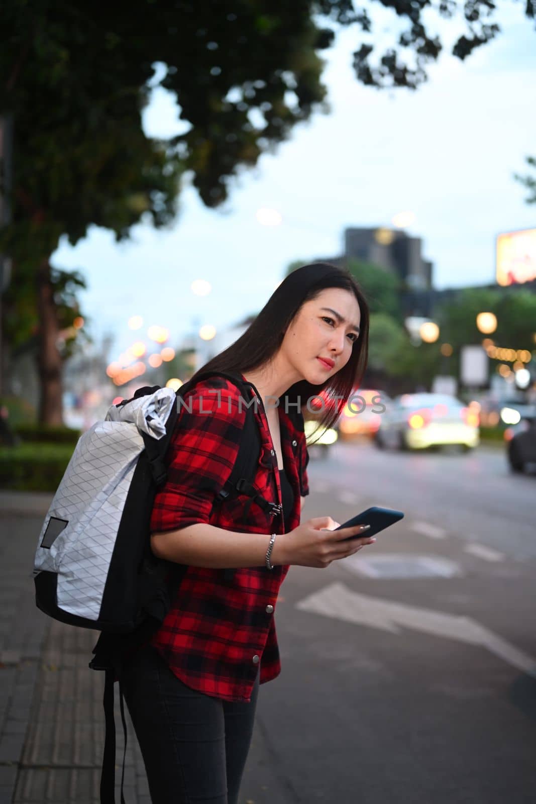Young Asian woman standing near the road with a smartphone in her hand and waiting for a taxi.