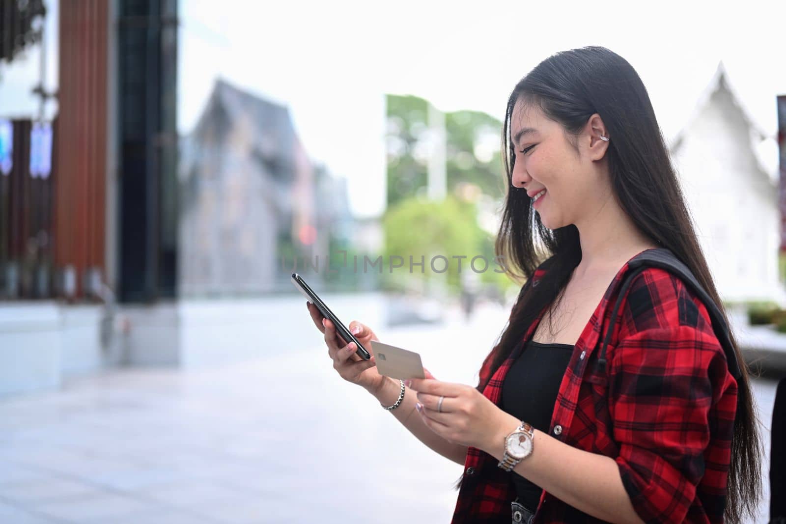 Young woman holding credit card and using smart phone for banking online while sitting at outdoor. by prathanchorruangsak