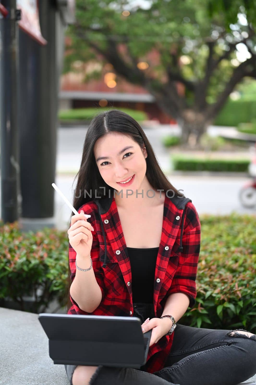 Smiling young woman holding digital tablet and smiling to camera while sitting at outdoor. by prathanchorruangsak