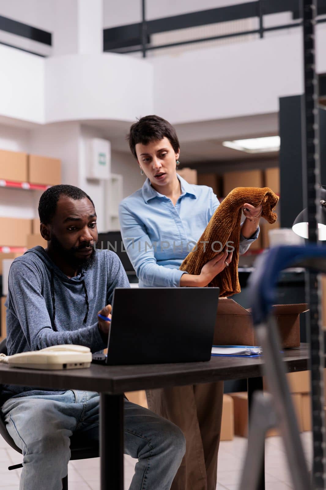 Fashion storehouse manager putting trendy blouse in carton box while supervisor man analyzing distribution on laptop by DCStudio
