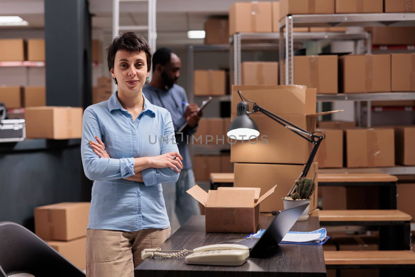 Warehouse manager standing in delivery department with arm crossed after finishing packing clients orders preparing packages for shipping. Diverse team working distribution center fulfillment company