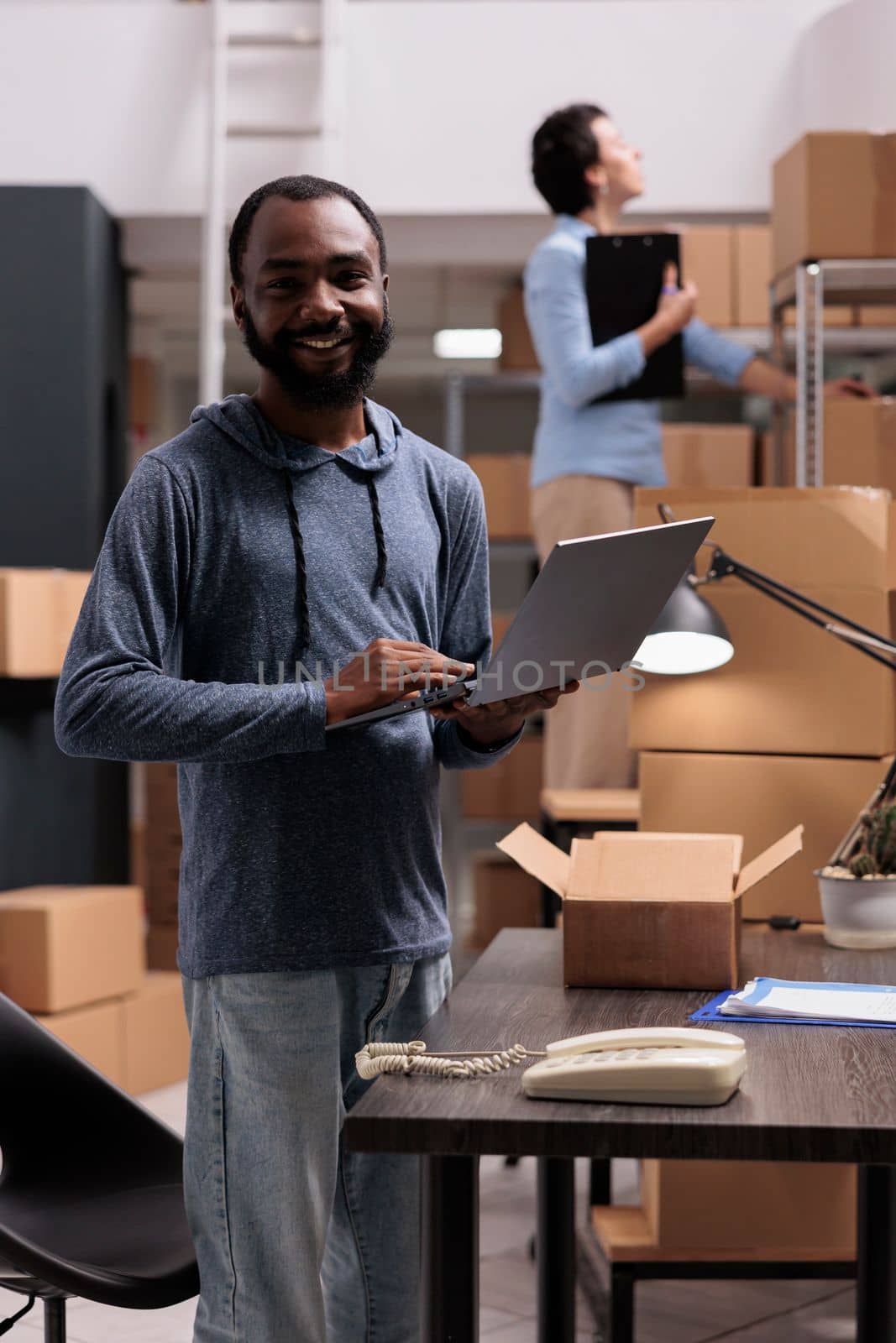 Smiling african american employee holding laptop computer looking at cargo stock working in warehouse delivery department. Storehouse employees preparing packages for customers orders
