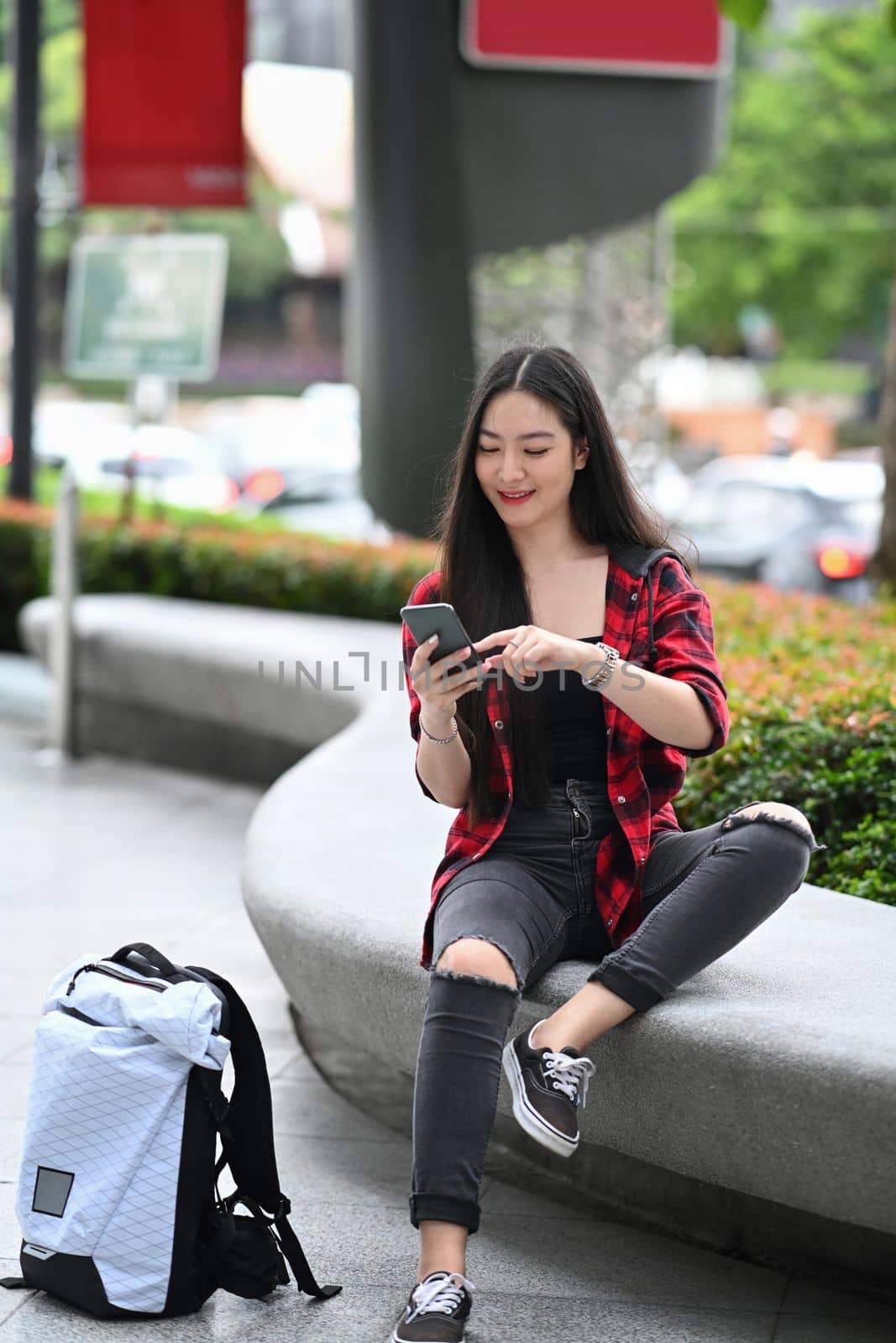 Smiling young Asian woman sitting in the city and using smart phone. by prathanchorruangsak