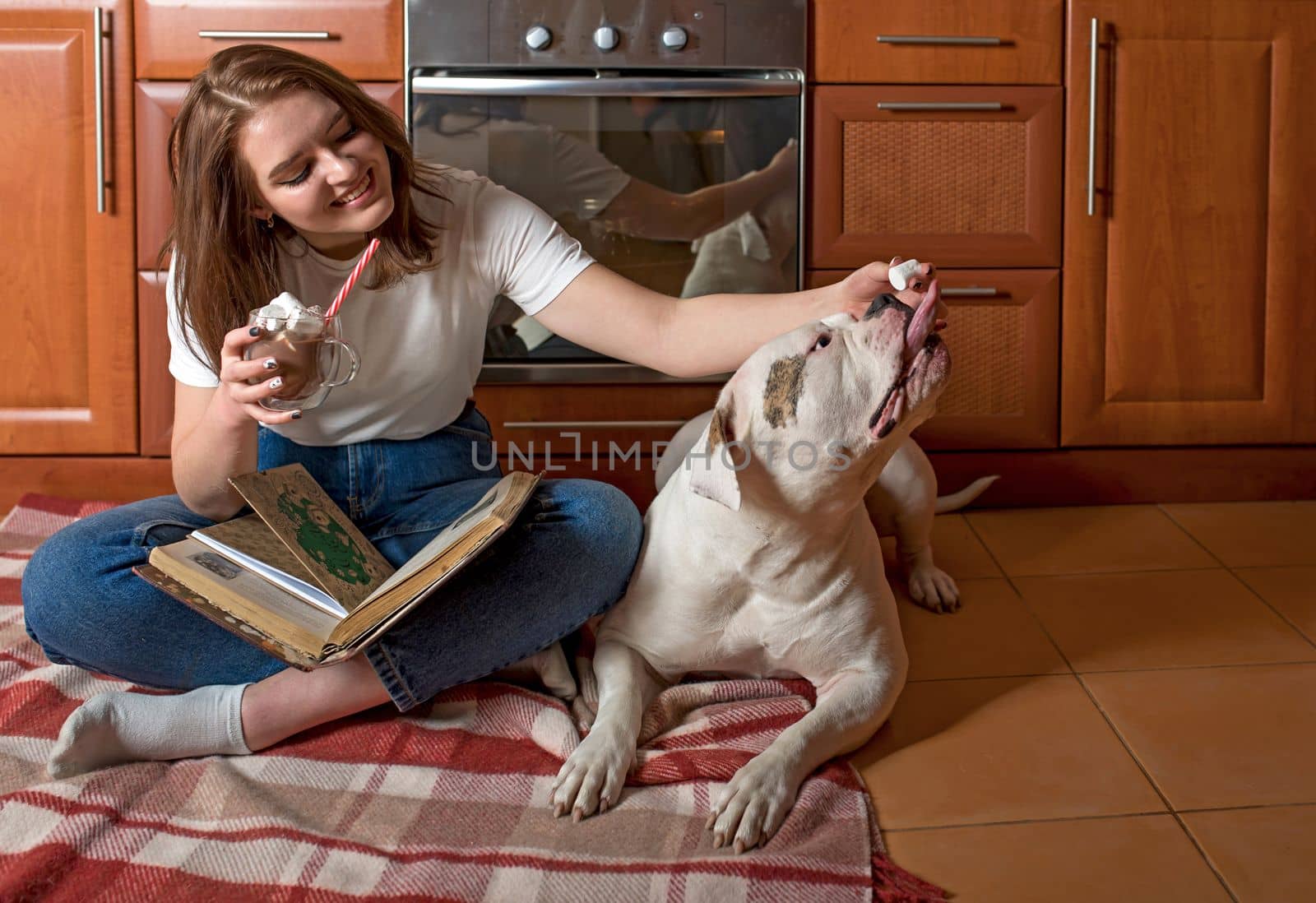 Happy girl, sitting on the floor in the kitchen, pint cocoa and playing with the dog by aprilphoto