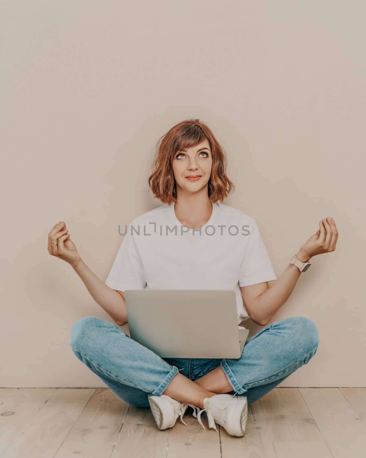 A brunette sits on the floor with a laptop on a beige wall background. She is wearing a white T-shirt, jeans and white sneakers
