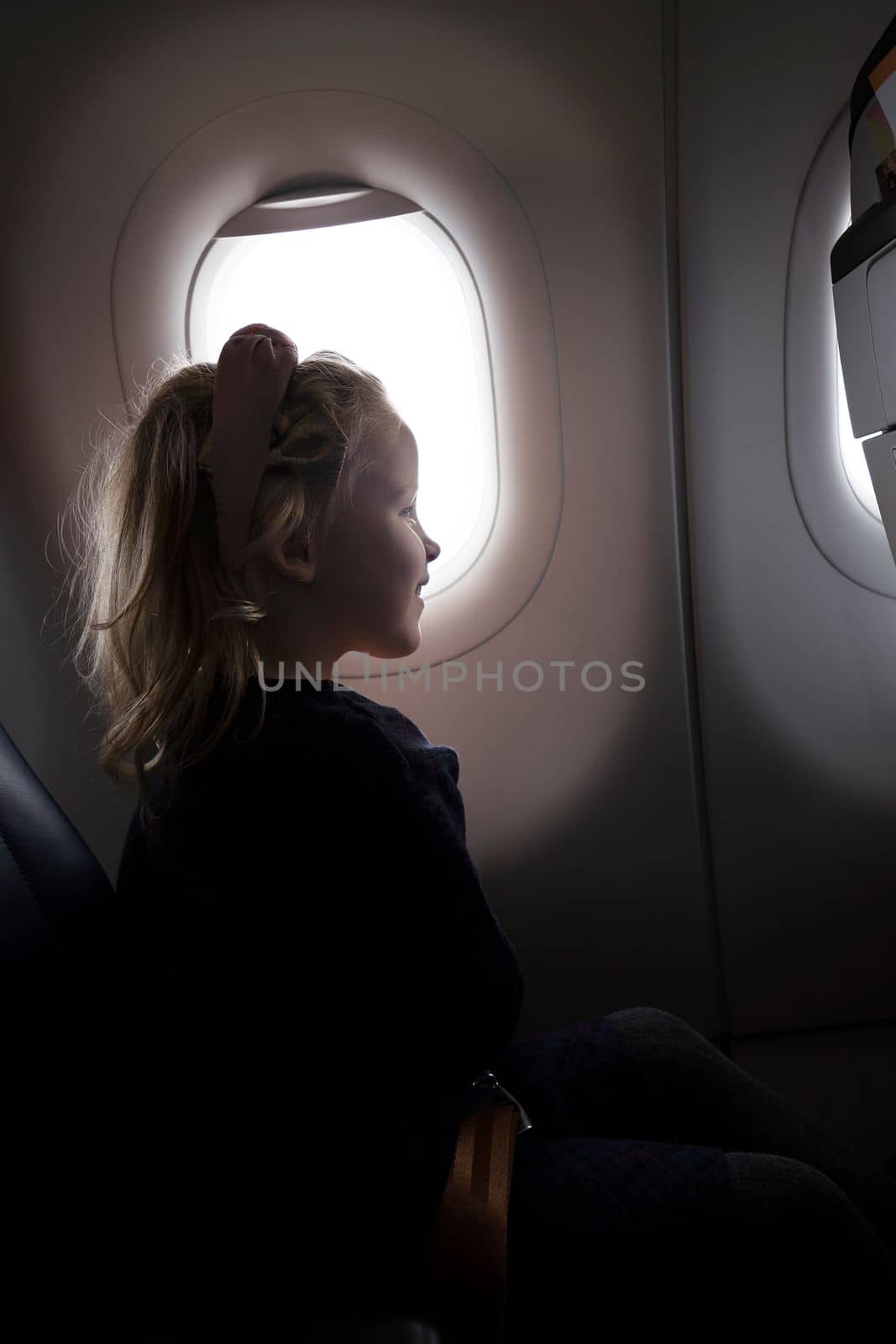 Side view of smiling preteen girl in dress sitting on passenger seat in aircraft near porthole