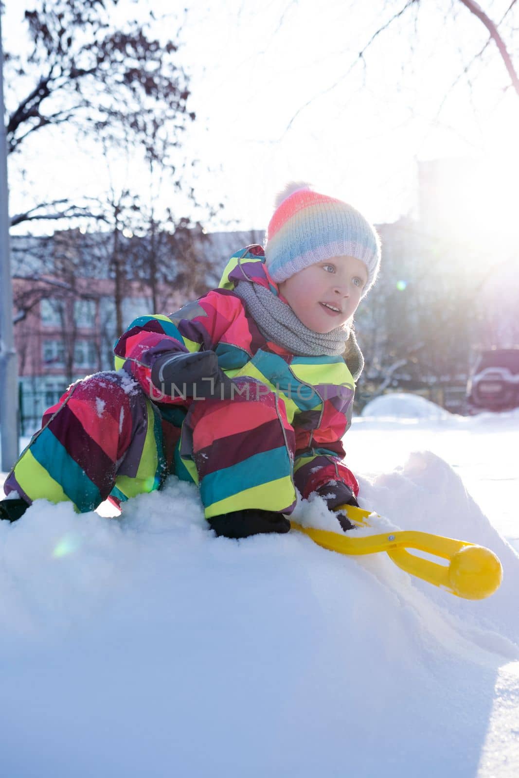 Full body of positive preteen girl in colorful warm clothes sitting on snowy hill with toy for making snowballs while playing in yard
