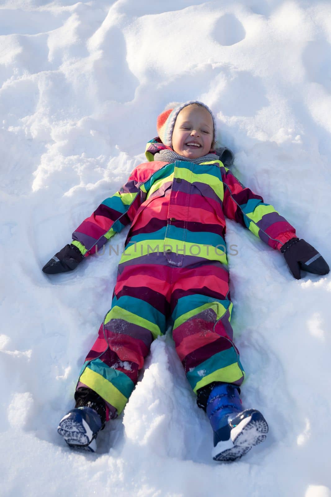 From above full body of laughing preteen girl in colorful outerwear making snow angel while playing outdoors on sunny winter day