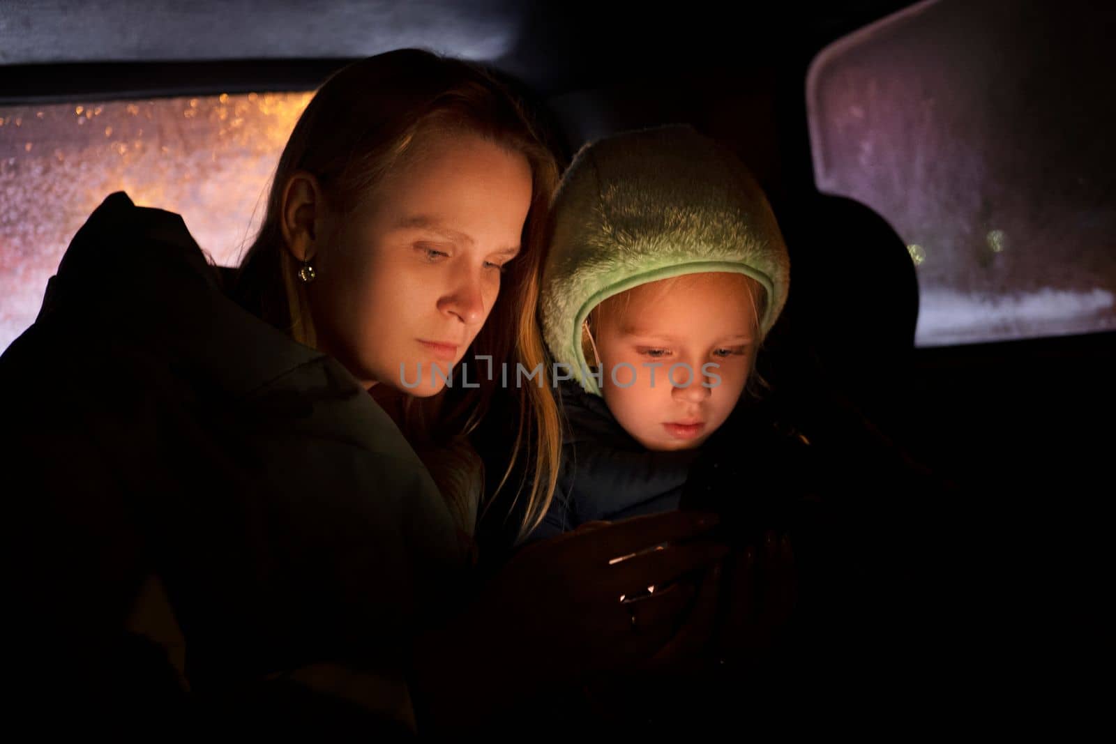 Serious woman in outerwear sitting in car with little daughter in hat and using smartphone during night trip