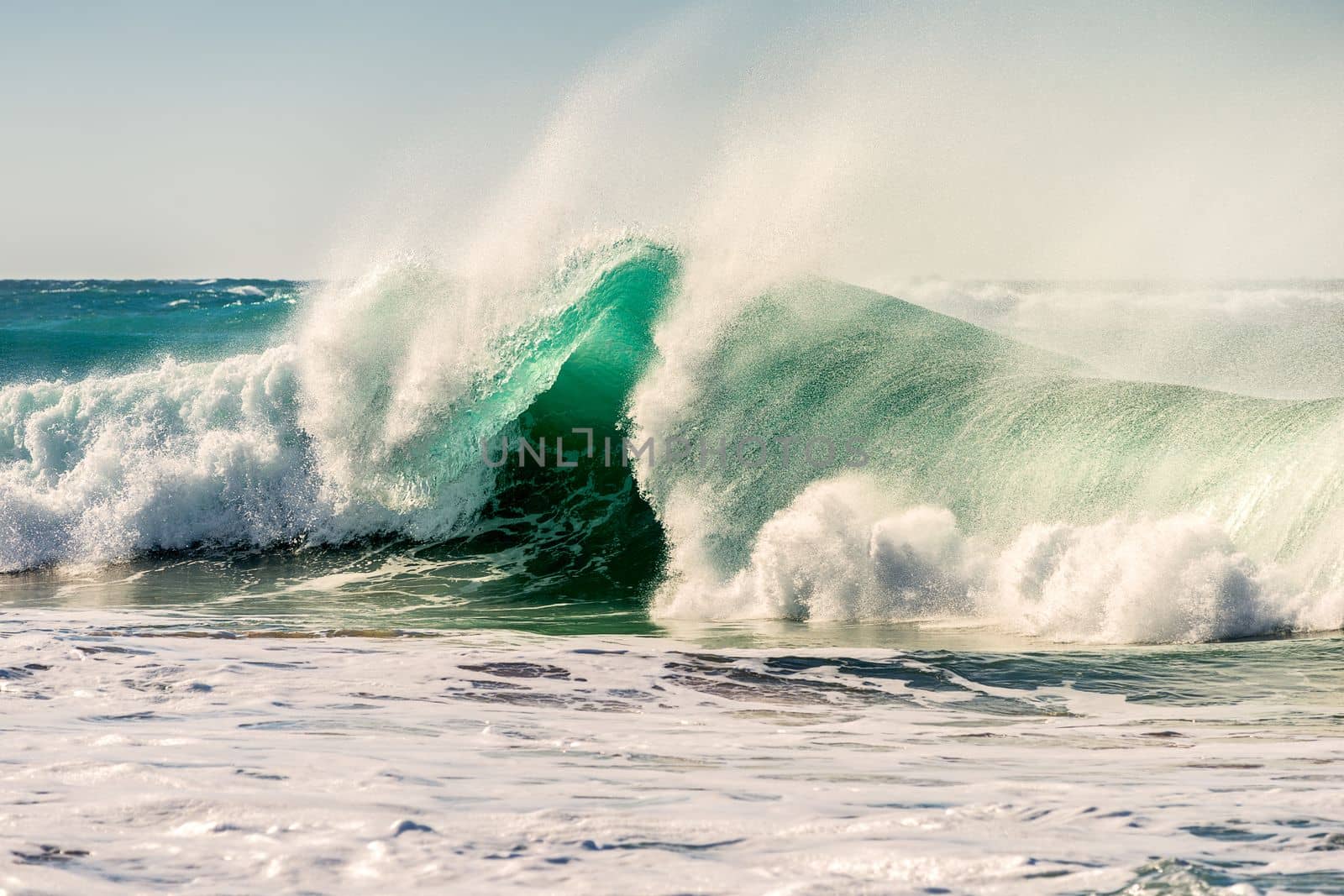 powerful wave breaking on the shore between foam and large plumes of vaporized water, the sun shines on the blue and turquoise surface of the water, the sky is clean and clear