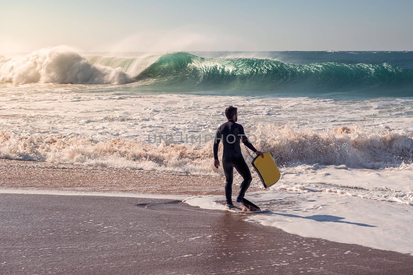 Man on the seashore prepares to surf, wears black neoprene wetsuit, in his hand has a yellow bodyboard. An awesome wave breaks with lots of energy and power, sunlight reflects on the huge pile of foam